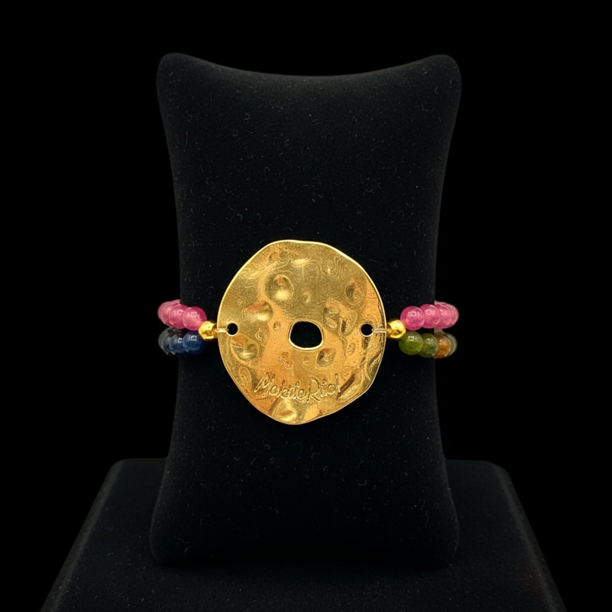 Pulsera elástica con piedras naturales y pieza dorada con logo de Maldita Rita joyería, en expositor negro