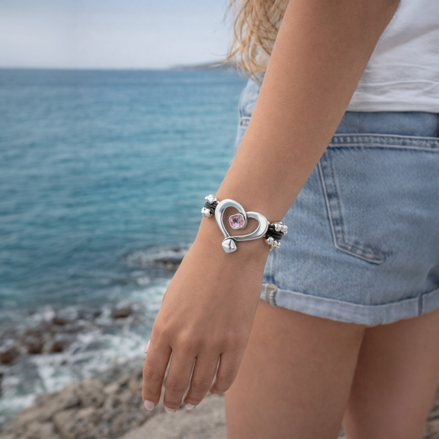 Pulsera de cuero y bolasde plata con corazón hueco y cristal de Swarovki rosa, de la marca Maldita Rita. Modelo en la playa.